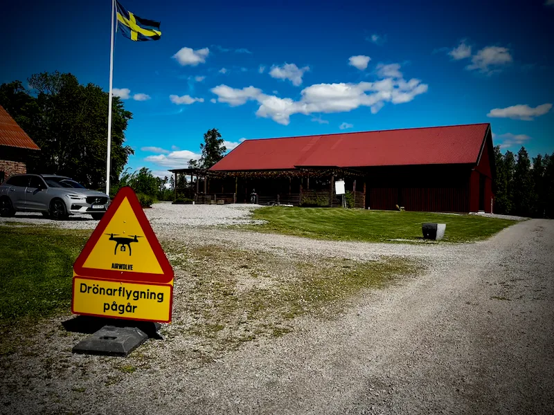 Varningsskylt “Drönarflygning pågår” uppställd vid en gård med röd lada